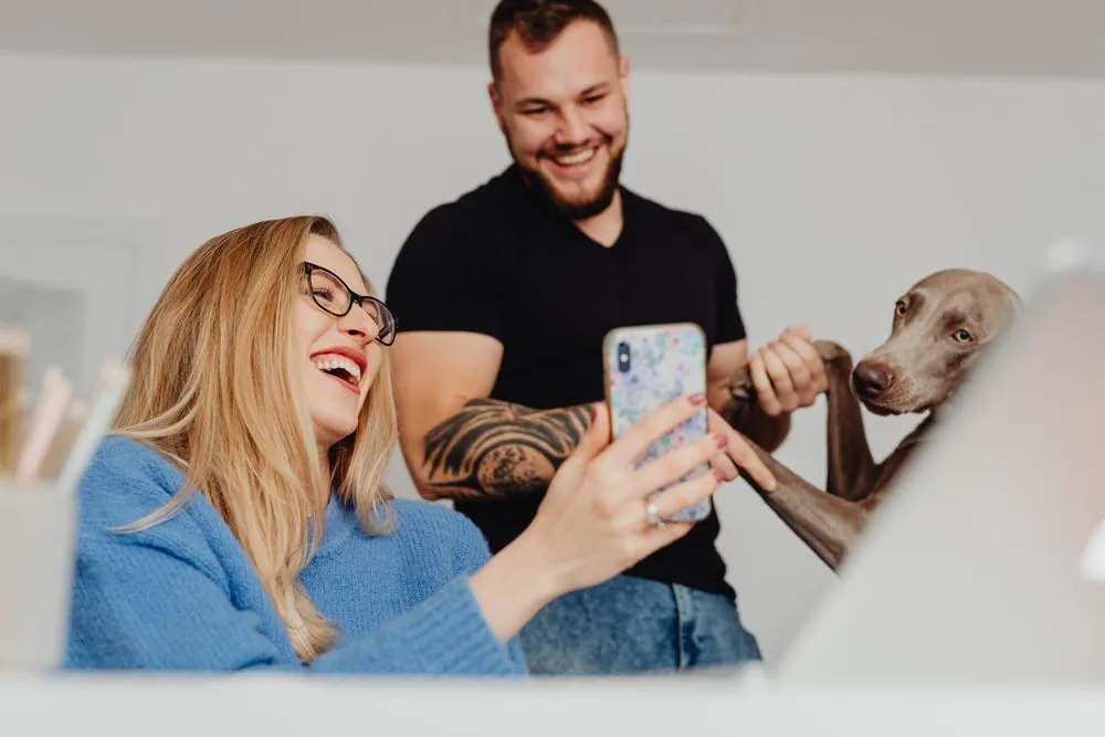 Pareja riéndose frente al celular junto a su perro mientras comparten la invitación por WhatsApp